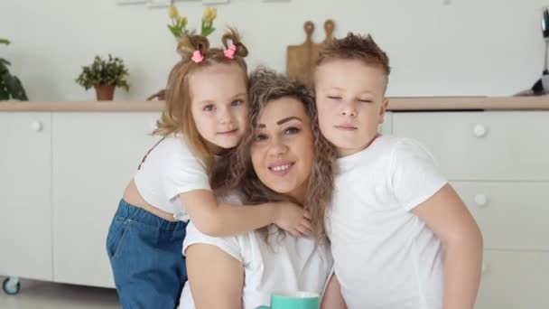 Famille amicale à la maison. Un garçon, une fille et une mère s'assoient par terre et s'embrassent dans une cuisine moderne blanche élégante au design scandinave et regardent la caméra. Famille en T-shirts et jeans blancs — Video