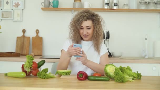 Mujer rubia joven con el pelo rizado hace una foto de verduras en un teléfono inteligente, con una cocina blanca en el fondo. Verduras frescas están sobre la mesa — Vídeos de Stock