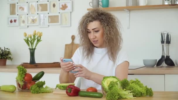 Mujer rubia joven con el pelo rizado toma fotos de verduras con un teléfono inteligente sentado en una mesa en la cocina diseñada en un estilo escandinavo Hugge — Vídeos de Stock