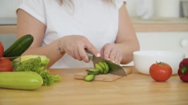 Kıvırcık saçlı genç sarışın bir kadın, İskandinav tasarım birliğiyle mutfaktaki bir masada otururken bir salatalığı yüzüklere ayırıyor. El hareketlerine yakından bak. Sağlıklı gıda ve diyet konsepti