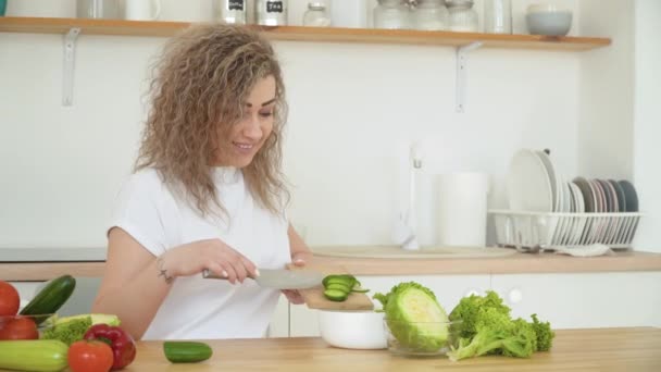 Jeune femme blonde aux cheveux bouclés jette un concombre tranché de la planche dans un bol tout en étant assis à une table dans la cuisine dans un design scandinave — Video