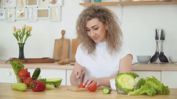 Giovane donna bionda con i capelli ricci taglia un pomodoro rosso seduto a un tavolo in cucina in un design scandinavo Hygge. Cibo sano e concetto di dieta — Video Stock