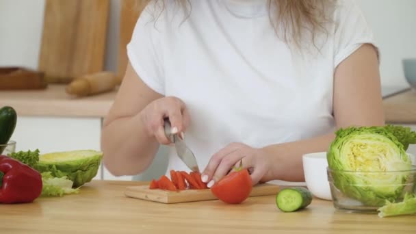 Молодая блондинка с вьющимися волосами режет красный помидор, сидя за столом на кухне. Крупный план ножа в руках — стоковое видео