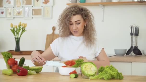 Kıvırcık saçlı genç sarışın kadın sıcak, parlak mutfaktaki masada otururken dilimlenmiş kırmızı domatesi kaseye atıyor. Sağlıklı gıda ve diyet konsepti — Stok video