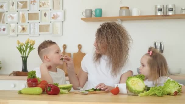 Junge blonde Frau mit lockigem Haar gibt ihrem Sohn und ihrer Tochter, die an einem Tisch in der Küche sitzen, einen Vorgeschmack auf ein Stück Gurke. Das Mädchen weigert sich, Gemüse zu essen — Stockvideo