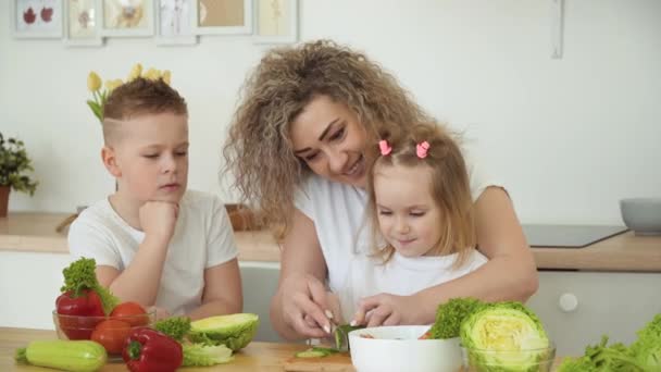 De kinderen bereiden samen met hun moeder een salade aan tafel in de keuken. De familie is gekleed in dezelfde basic witte T-shirts — Stockvideo