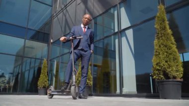 Klasik mavi takım elbiseli bir Afro-Amerikalı elinde elektrikli scooterıyla modern bir ayna binasının yanında duruyor. Alt görünüm tam uzunlukta