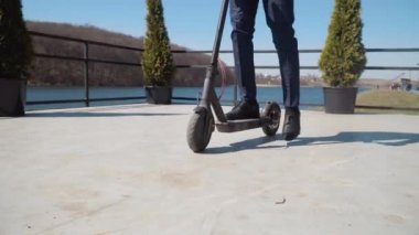 Klasik takım elbiseli ve mavi ayakkabılı bir adam nehir kıyısında elektrikli scooter kullanıyor. Alt görünüm tam uzunlukta