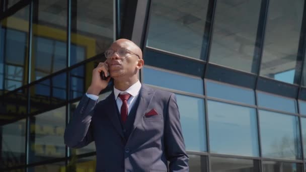 Fiatal, vonzó afro-amerikai üzletember beszél telefonon egy modern tükörépület közelében. Okostelefon, szörfözés. Modern technológia és kommunikáció — Stock videók
