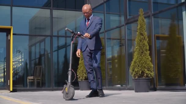 Un hombre afroamericano con un traje de negocios azul clásico se sube a un scooter eléctrico y se pasea por la calle cerca de un moderno edificio espejo. Vista inferior en longitud completa — Vídeo de stock