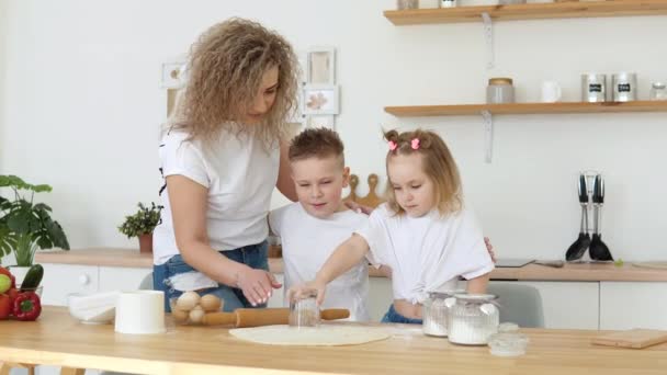 Οικογενειακή διασκέδαση με παιδιά. Δημιουργικότητα και μαγειρική για παιδιά. Μητρότητα. Μια οικογένεια ξανθών με λευκά μπλουζάκια έκοψε μπισκότα από ζύμη σε μια λευκή σκανδιναβική κουζίνα. — Αρχείο Βίντεο