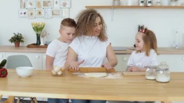 Beyaz tişörtlü beyaz sarışınların ailesi. Bir anne, oğlu ve kızıyla birlikte İskandinav Hygge tasarımı ile mutfakta hamur yoğurur ve yuvarlar.