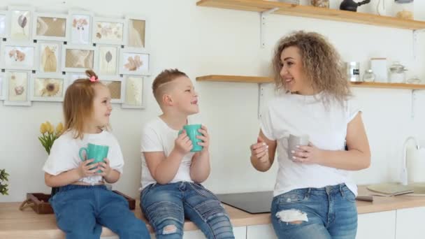 Хлопчик, дівчинка і мати п'ють напій з синіх чашок, сидячи на столі в стильній білій сучасній кухні в скандинавському дизайні. Родина в білих футболках і джинсах — стокове відео