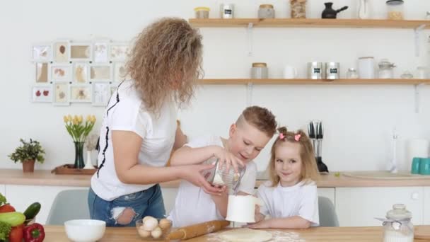 Le garçon verse la farine dans un tamis quand lui et sa mère et sa sœur déploient la pâte dans la cuisine. Une famille de blondes en T-shirts blancs — Video