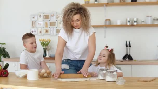 Oğlan ve kız, anne mutfakta hamuru yuvarlarken onu izliyorlar. Beyaz tişörtlü sarışınlardan oluşan bir aile ortaya çıkıp hamuru yoğuruyor. — Stok video