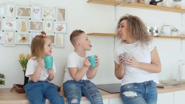 Chico, niña y madre bromean y beben una copa de tazas azules sentadas en la mesa en una elegante cocina moderna blanca en diseño escandinavo. Familia en camisetas blancas y jeans — Vídeos de Stock