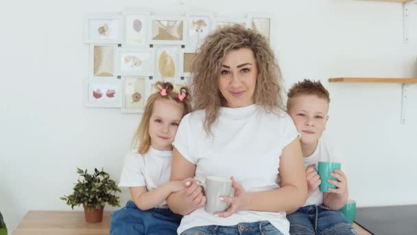 一个男孩、一个女孩和一个母亲坐在斯堪的纳维亚风格时尚的白色现代厨房的桌子旁，看着摄像机。穿着白色T恤衫和牛仔裤的家庭 — 图库视频影像