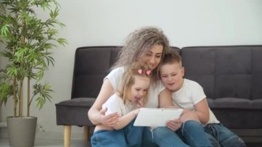 Anne, kız ve oğul sıcak bir oturma odasında tabletle oynuyorlar. Ailede de aynı beyaz tişörtler var. Çocuklarla vakit geçirmek. Aile eğlencesi