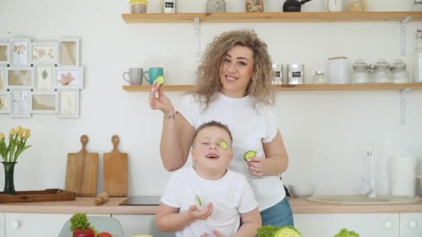 Η μαμά και ο γιος κόβουν λαχανικά μαζί και παίζουν μαζί τους. Φρέσκα λαχανικά είναι στο τραπέζι. Υγιεινή διατροφή. Παιδικό μενού. Ικανότητες αυτοφροντίδας. Η οικογένεια είναι ντυμένη με τα ίδια βασικά λευκά μπλουζάκια. — Αρχείο Βίντεο