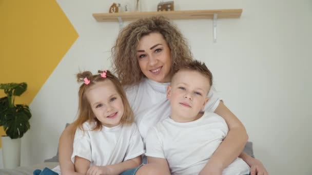 Filha e filho sentam-se nos braços das mães e beijam as bochechas das mães. Família em um quarto acolhedor. Belo retrato de família. Cores branco e amarelo. Minimalismo — Vídeo de Stock