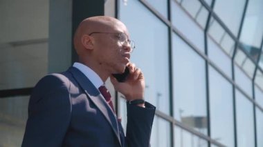 Genç ve çekici Afrikalı Amerikalı iş adamı modern bir ayna binasının yanında telefonla konuşuyor. Akıllı telefon tutmak, ağlarda sörf yapmak. Modern teknoloji ve iletişim