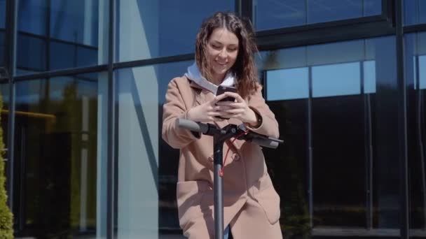 Süveter, kot pantolon ve palto giyen, modern bir binanın yanında elektrikli scooter kullanan ve akıllı telefon kullanan genç bir öğrenci. Gençler ve iş adamları için modern aletler — Stok video