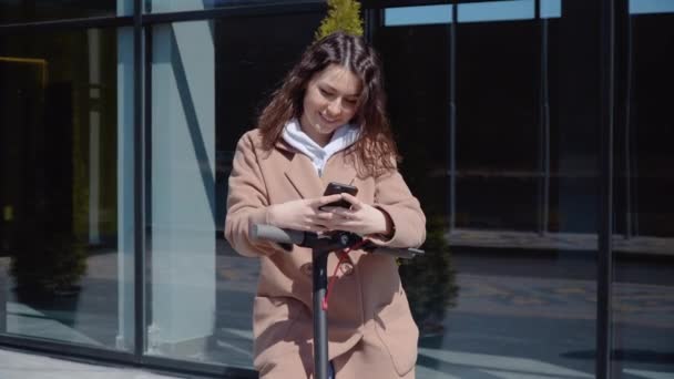 Süveter, kot pantolon ve palto giyen, modern bir binanın yanında elektrikli scooter kullanan ve akıllı telefon kullanan genç bir öğrenci. Ön görünüm — Stok video