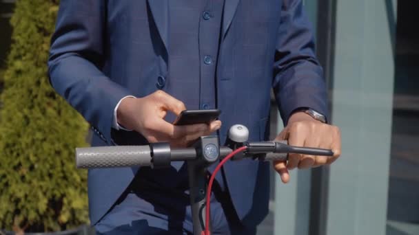 Un uomo afro-americano in un classico abito da lavoro blu si erge con uno scooter elettrico per strada vicino a un moderno edificio a specchio e utilizza uno smartphone. Tipo di mani e volante — Video Stock