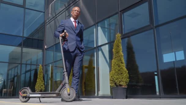 Un afroamericano in un classico abito da lavoro blu si erge con uno scooter elettrico per strada vicino a un moderno edificio a specchio con la mano in tasca. Vista in basso a tutta lunghezza — Video Stock