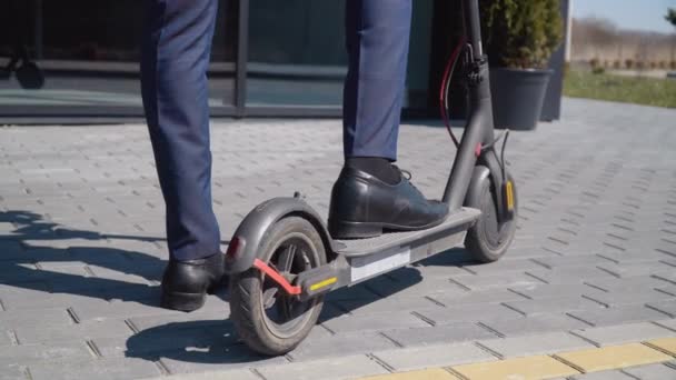 Un hombre afroamericano con un traje de negocios clásico y zapatos azules está de pie con un pie en una scooter eléctrica cerca de un edificio de espejo moderno. Vista inferior en longitud completa — Vídeo de stock