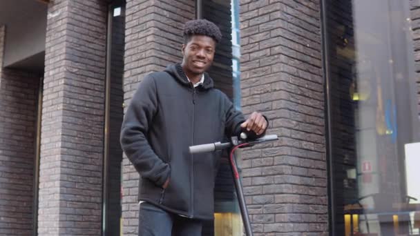 Een jonge Afro-Amerikaanse man in casual donkere kleren met een scooter staat in de buurt van een gebouw met een donkere bakstenen gevel — Stockvideo