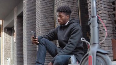 Siyah kazaklı, siyah kot pantolonlu ve spor ayakkabılı genç bir Afrikalı Amerikalı siyah tuğlalı bir yüzle kaldırımda oturuyor. Etkin bir yaşam tarzı ve iletişim için araçlar