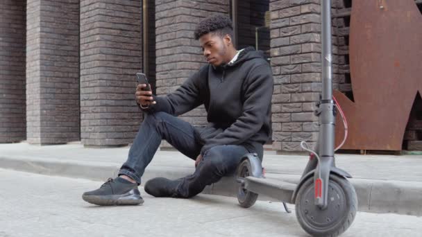 黒いセーターを着た若いアフリカ系アメリカ人の男性と黒いジーンズとスニーカーが暗いレンガ造りのファサードのある建物の近くの歩道に座っています。若者の都市生活 — ストック動画