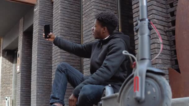 Un joven afroamericano con pantalones vaqueros y zapatillas negras se sienta en la acera cerca de un edificio con una fachada de ladrillo oscuro. El tipo se toma una selfie con un scooter eléctrico — Vídeo de stock