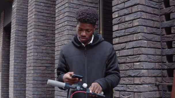 Um jovem afro-americano em roupas escuras casuais com uma scooter fica perto de um edifício com uma fachada de tijolo escuro e usa um smartphone. Retrato na cintura — Vídeo de Stock
