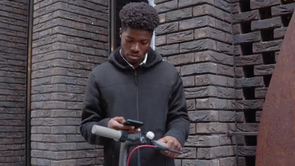 Um jovem afro-americano em roupas escuras casuais com uma scooter fica perto de um edifício com uma fachada de tijolo escuro e tipos em um smartphone. Retrato na cintura — Vídeo de Stock