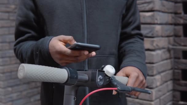 A young African-American in casual dark clothes with a scooter stands near a building with a dark brick facade and types on a smartphone. Close-up view of hands and steering wheel — Stock Video