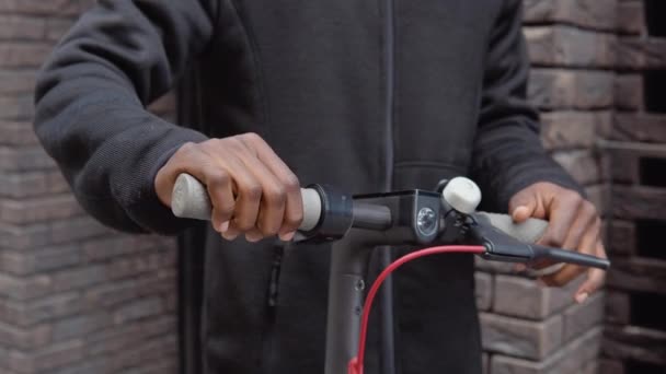 Un joven afroamericano en ropa oscura casual con una scooter se encuentra cerca de un edificio con una fachada de ladrillo oscuro y tipos en un teléfono inteligente. Vista de cerca de las manos y el volante — Vídeos de Stock
