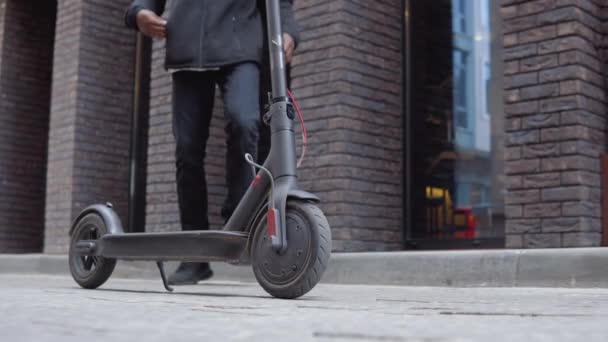 Ein junger afroamerikanischer Mann in schwarzem Pullover, schwarzen Jeans und Turnschuhen nähert sich einem Elektroroller und fährt los. Blick auf die Füße und die Straße von unten — Stockvideo