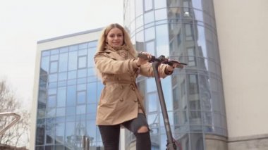 Bej renkli trençkotlu ve kotlu aktif genç şık sarışın kız modern bir gökdelenin arka planında elektrikli scooterla duruyor.