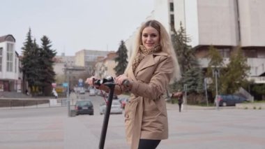 Bej renkli trençkotlu ve kot pantolonlu şık sarışın kız arka plandaki rahat sokakta elektrikli scooter ile duruyor.