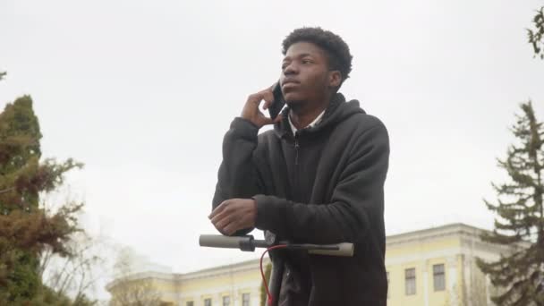 Egy fiatal afro-amerikai férfi fekete pulóverben, fekete farmerban és tornacipőben rátámaszkodik egy elektromos robogóra és mobilon beszél. — Stock videók