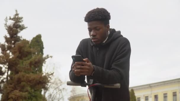 Um jovem afro-americano em uma camisola preta e jeans e tênis pretos se inclina em uma scooter elétrica e olha em um telefone celular — Vídeo de Stock