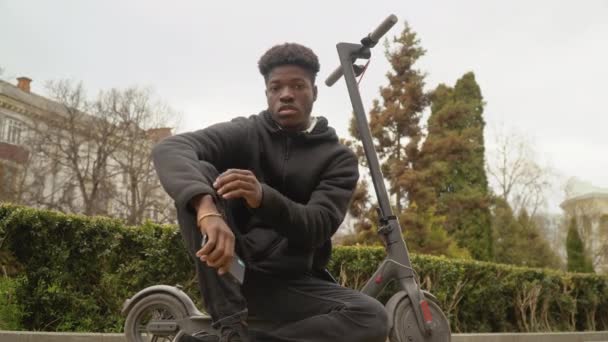 Een jonge Afro-Amerikaanse man in een zwarte trui en zwarte jeans en sneakers zit op een elektrische scooter in de buurt van het gazon en kijkt naar de camera met een smartphone in zijn handen. — Stockvideo