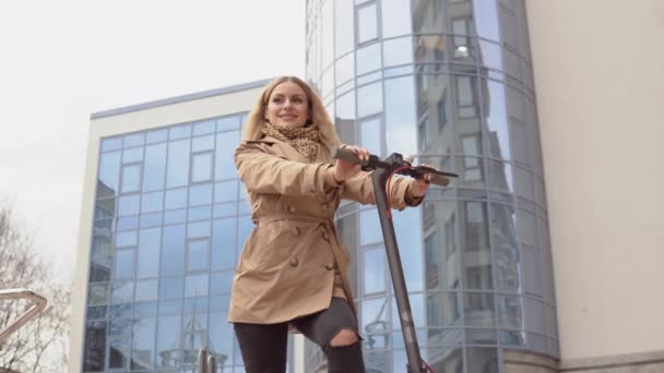 Joven chica rubia con estilo en un abrigo de trinchera beige y pantalones vaqueros con una scooter eléctrico en el fondo de un moderno rascacielos — Vídeos de Stock