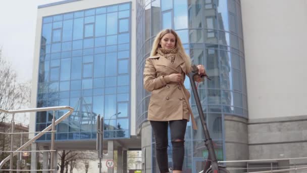 Joven chica rubia con estilo en un abrigo de trinchera beige y pantalones vaqueros con una scooter eléctrico en el fondo de un moderno rascacielos — Vídeos de Stock