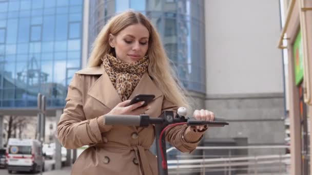 Joven mujer rubia con estilo en un abrigo de trinchera beige y jeans toma scooter patada eléctrica en compartir estacionamiento. Aplicación de teléfono turístico — Vídeo de stock