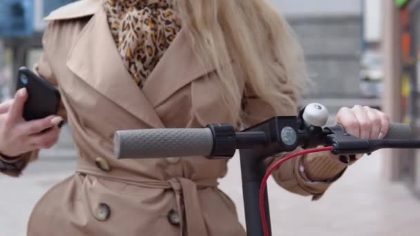 Una mujer joven alquila scooter eléctrico utilizando la aplicación de teléfono móvil. Aplicación de teléfono turístico. Aparatos modernos para jóvenes y gente de negocios. Vista cercana de las manos, el teléfono y el volante — Vídeos de Stock