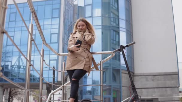 Молодая стильная блондинка в бежевом плаще и джинсах стоит на электрическом скутере с современным высотком на заднем плане и делает селфи — стоковое видео