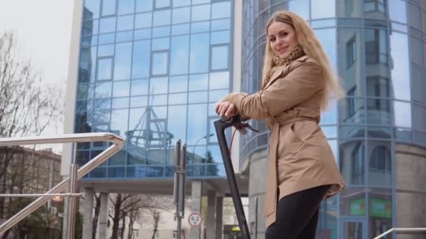 Jonge stijlvolle blonde meid in een beige trench jas en jeans staat met een elektrische scooter op de achtergrond van een moderne hoogbouw. Zijaanzicht — Stockvideo